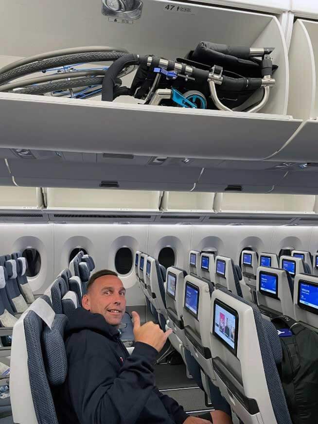 Shaun seated on an airplane with his wheelchair stored in the overhead compartment above him